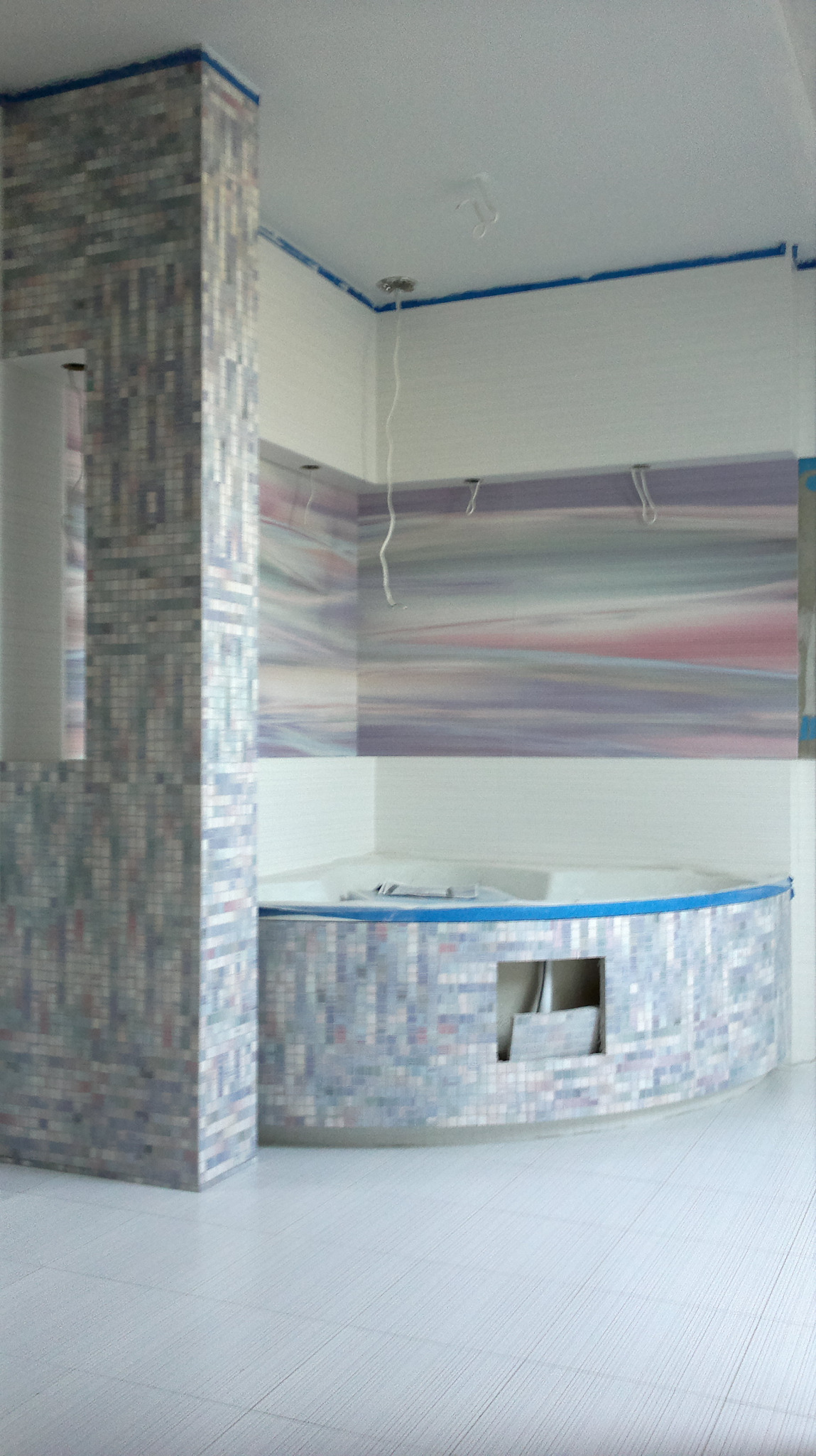 Niedokończona łazienka z mozaiką w odcieniach szarości, fioletu i błękitu na obudowie wanny i kolumnie, zabezpieczona niebieską taśmą malarską, widoczne wystające kable od oświetlenia.