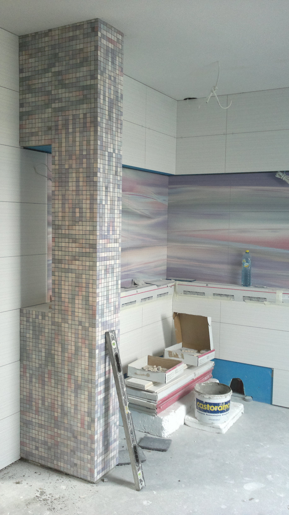 Wnętrze łazienki w trakcie prac wykończeniowych: kolumna obłożona mozaiką z drobnych płytek, poziomica oparta o ścianę, widoczne materiały budowlane i narzędzia w nieładzie.