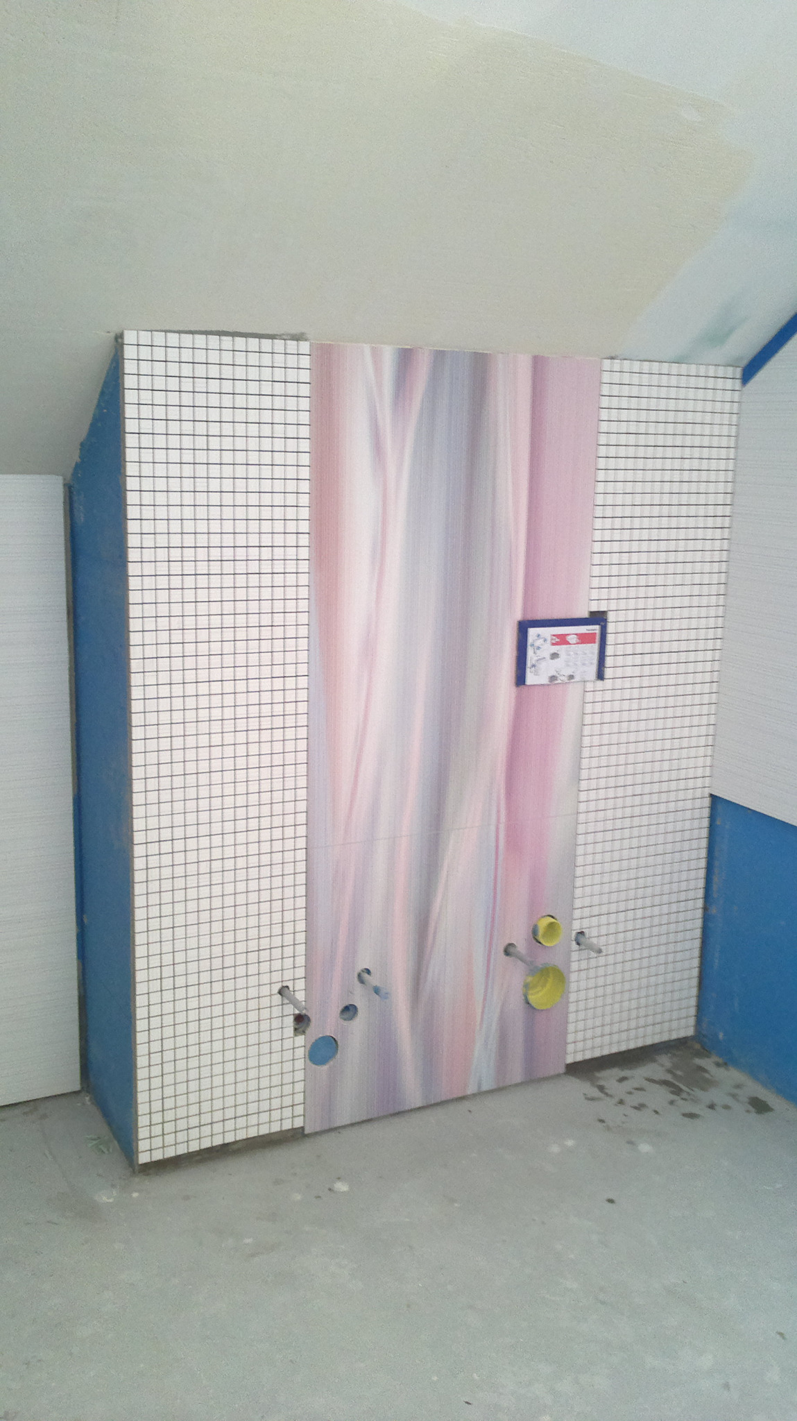 Ścianka działowa w łazience w trakcie remontu, obłożona mozaiką z białych płytek i panelami z abstrakcyjnym wzorem w odcieniach różu i fioletu, widoczne otwory na instalacje sanitarne.