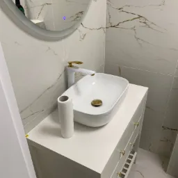 Nowoczesna łazienka w apartamencie: biała umywalka nablatowa z baterią w kolorze złota, marmurowe płytki na ścianach i podłodze, oraz okrągłe lustro z podświetleniem LED.