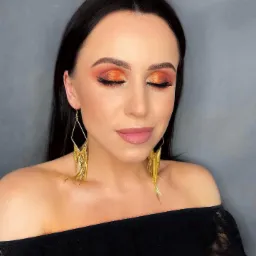 Kobieta z zamkniętymi oczami prezentuje makijaż oczu w odcieniach pomarańczy i złota, uzupełniony czarną kreską i sztucznymi rzęsami, nosząca długie, złote kolczyki w kształcie rombów z frędzlami.