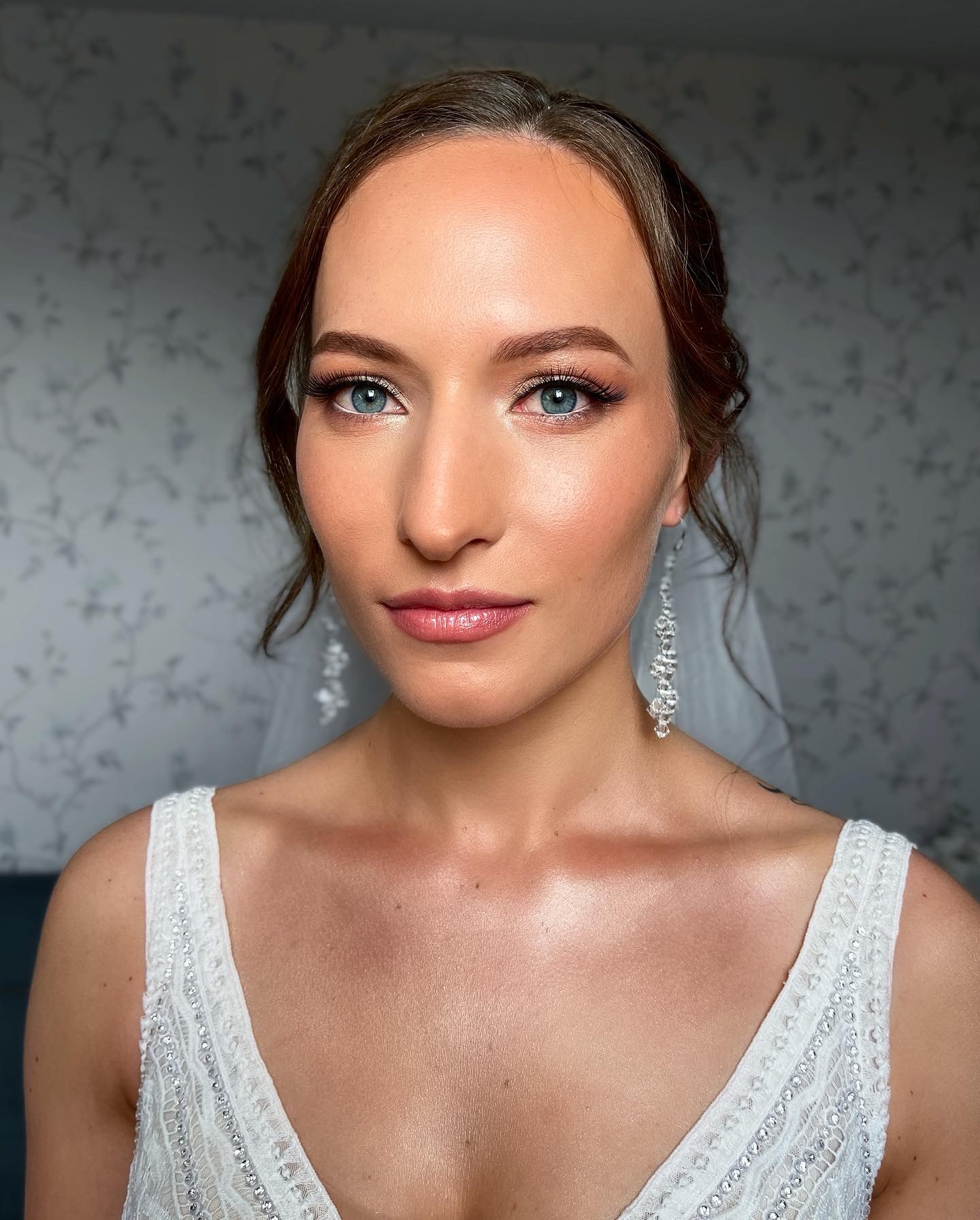 Portret kobiety z delikatnym, rozświetlonym upiększeniem ślubnym i długimi, wiszącymi kolczykami, ubrana w białą suknię z koronkowymi detalami na tle ściany w drobne kwiaty.