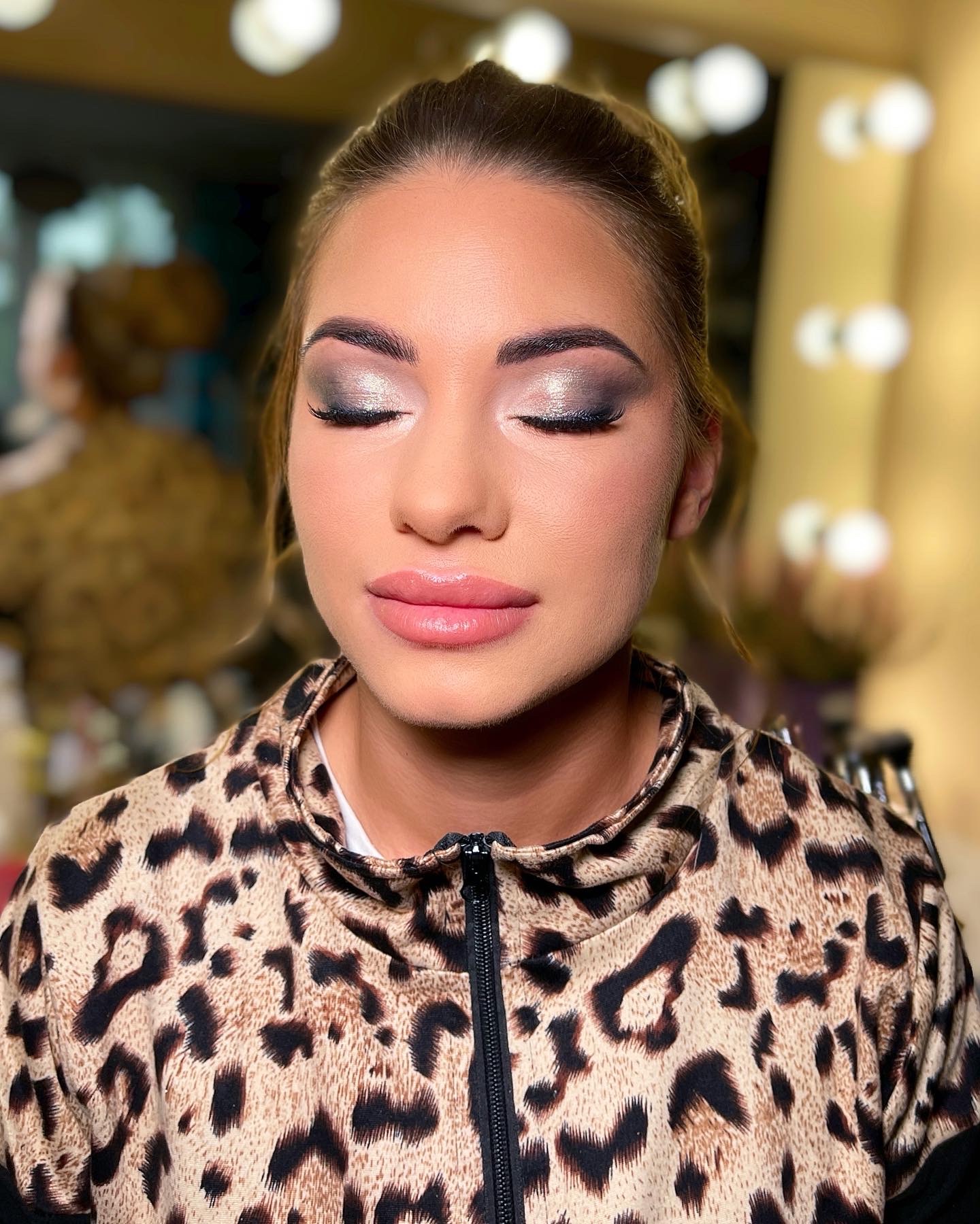 Portret kobiety z zamkniętymi oczami, prezentującej makijaż oczu w odcieniach szarości i srebra z błyszczącym akcentem, oraz delikatny makijaż ust z błyszczykiem, ubrana w bluzę w panterkę, na tle...