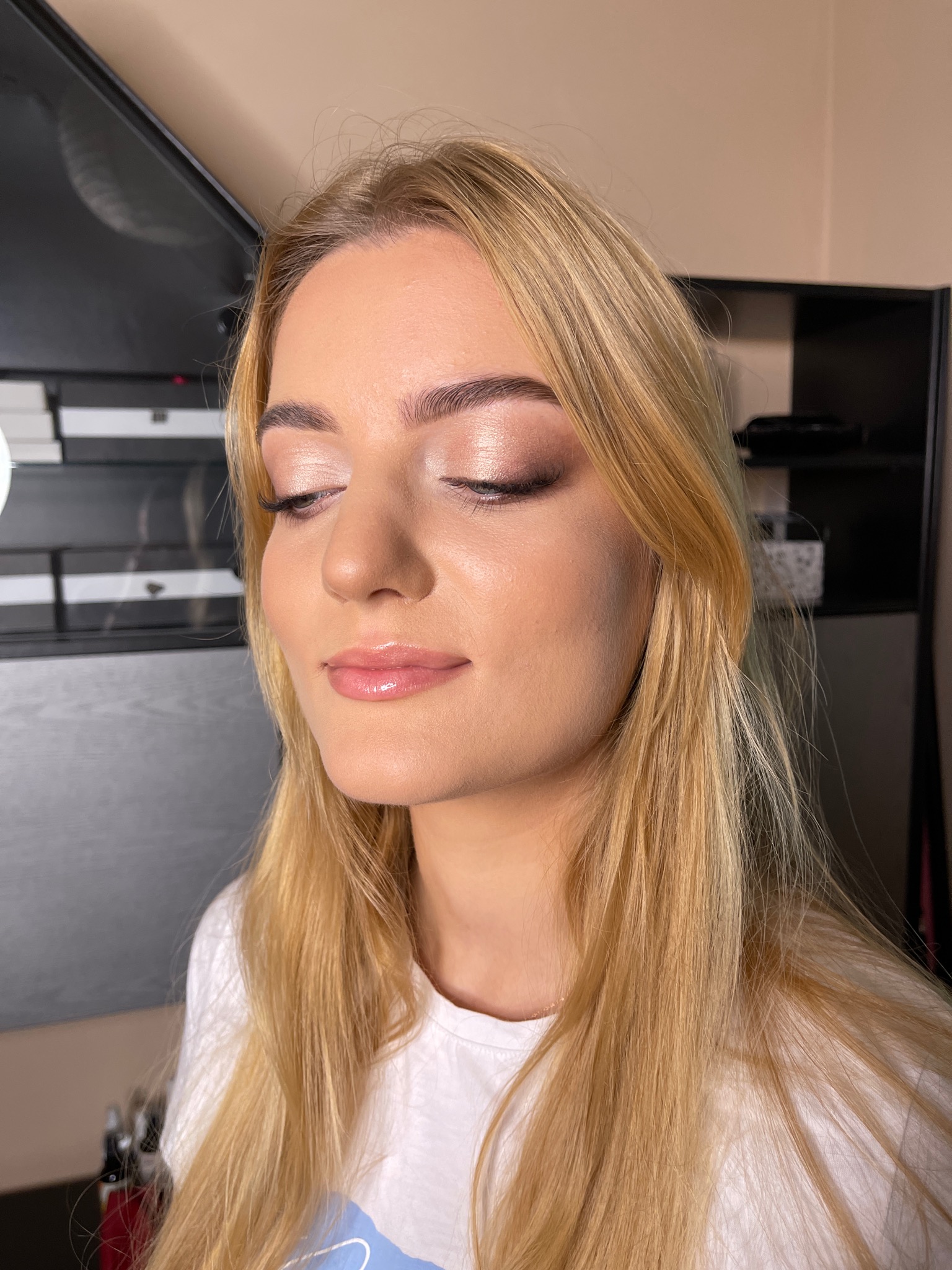 Portret kobiety z delikatnym, rozświetlonym makijażem oczu i błyszczącymi ustami, z zamkniętymi oczami i blond włosami opadającymi na ramiona, na neutralnym tle.