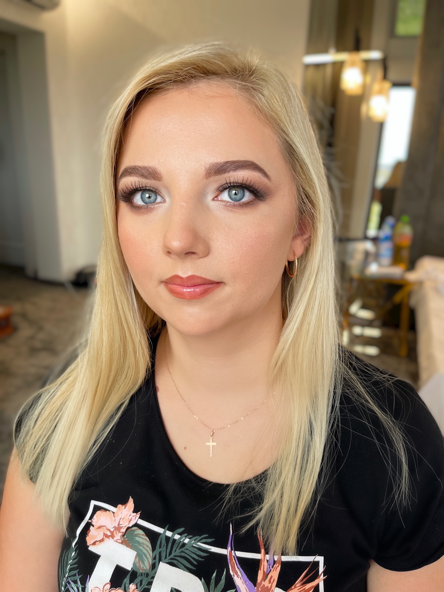 Portret kobiety z blond włosami i subtelnym, błyszczącym wykończeniem ust, podkreślonym spojrzeniem i złotą biżuterią na tle jasnego wnętrza.