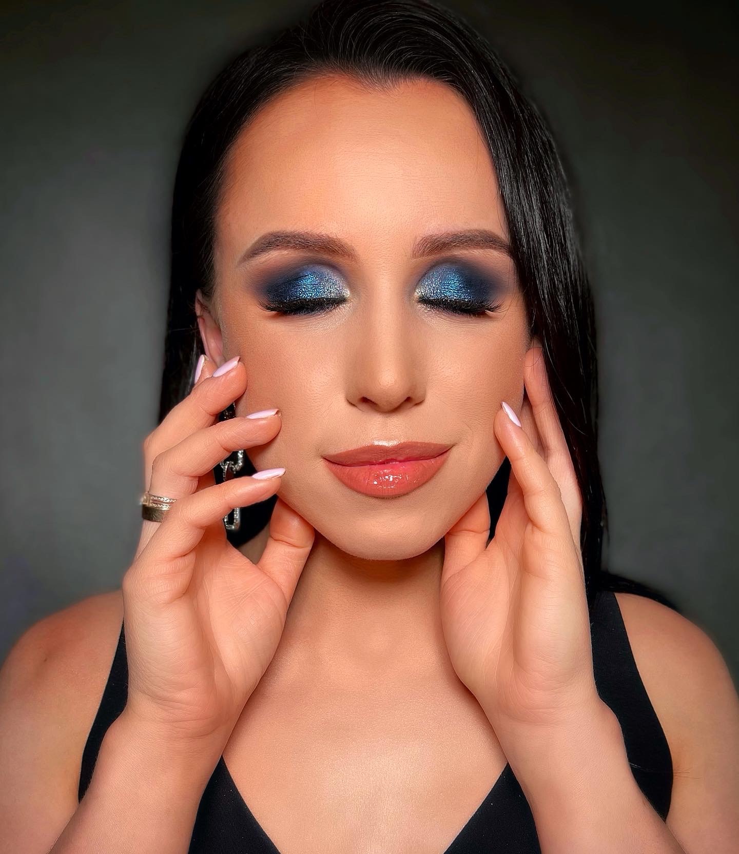 Portret kobiety z zamkniętymi oczami, prezentującej intensywny, brokatowy, granatowy cień na powiekach i błyszczące usta. Dłonie z delikatnym manicure obejmują twarz, eksponując pierścionek na palcu.