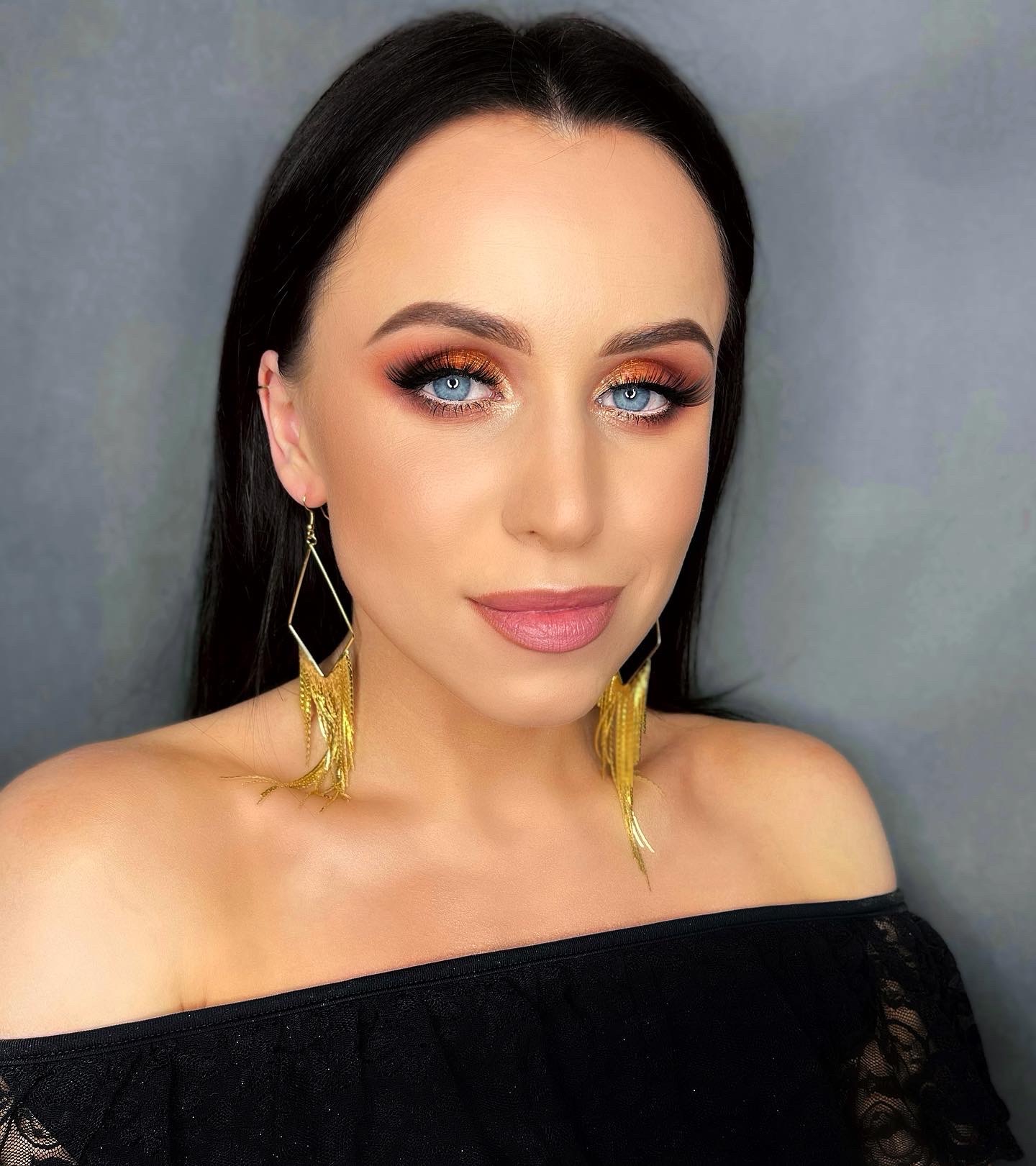 Portret kobiety z intensywnym, pomarańczowo-złotym cieniowaniem powiek, długimi sztucznymi rzęsami i delikatnym różem na ustach, uzupełniony dużymi, geometrycznymi kolczykami z frędzlami.