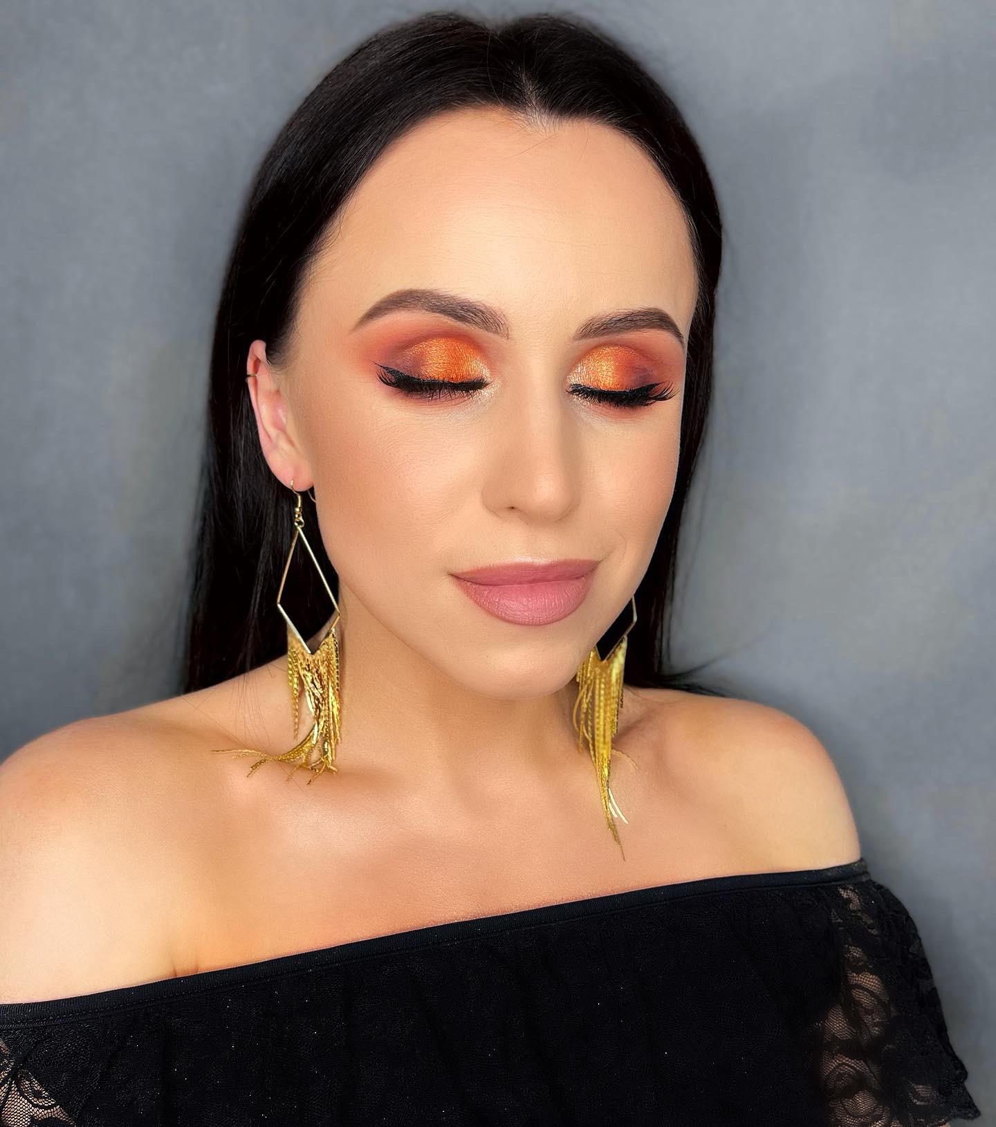 Kobieta z zamkniętymi oczami prezentuje makijaż oczu w odcieniach pomarańczy i złota, uzupełniony czarną kreską i sztucznymi rzęsami, nosząca długie, złote kolczyki w kształcie rombów z frędzlami.