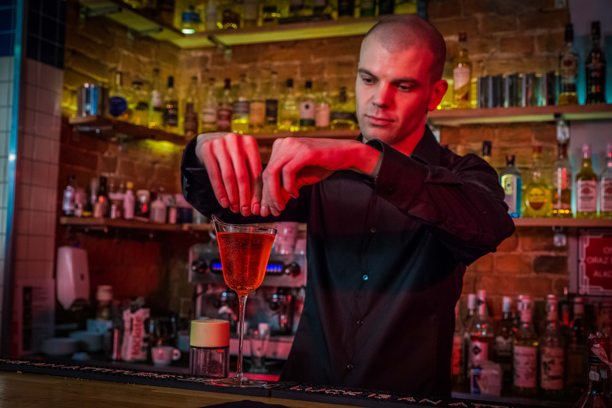 Barman w czarnej koszuli przygotowuje koktajl w kieliszku, wyciskając sok z pomarańczy nad barem z podświetlonymi butelkami w tle.