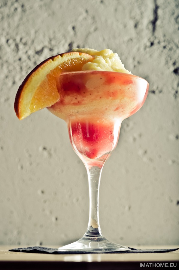 Szklanka z czerwono-pomarańczowym koktajlem i plasterkiem pomarańczy na krawędzi, stojąca na czarnej serwetce na tle szarej ściany.