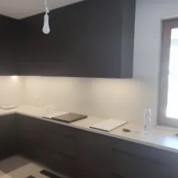 Częściowo zamontowana, nowoczesna kuchnia z ciemnymi szafkami, jasnym blatem i oświetleniem podszafkowym. Na blacie leżą próbki blatów, narzędzia i butelka wody. Widoczne niedokończone wykończenie...