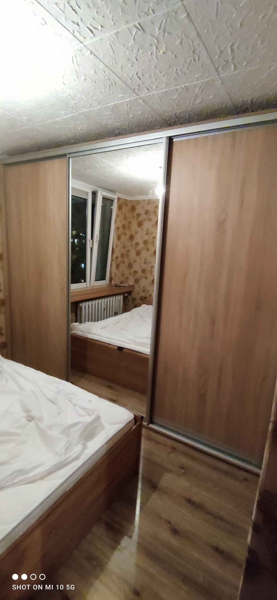 Szafa przesuwna z lustrem w sypialni, odbicie łóżka i okna w lustrze, widoczna faktura sufitu i wzór tapety.