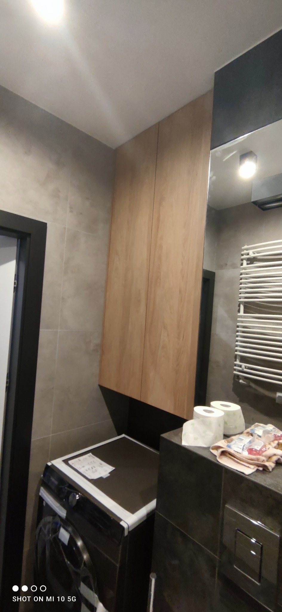 Wysoka, drewnopodobna szafka łazienkowa nad pralką, zamontowana na ścianie z szarych płytek, widoczny również fragment lustra i grzejnika drabinkowego.