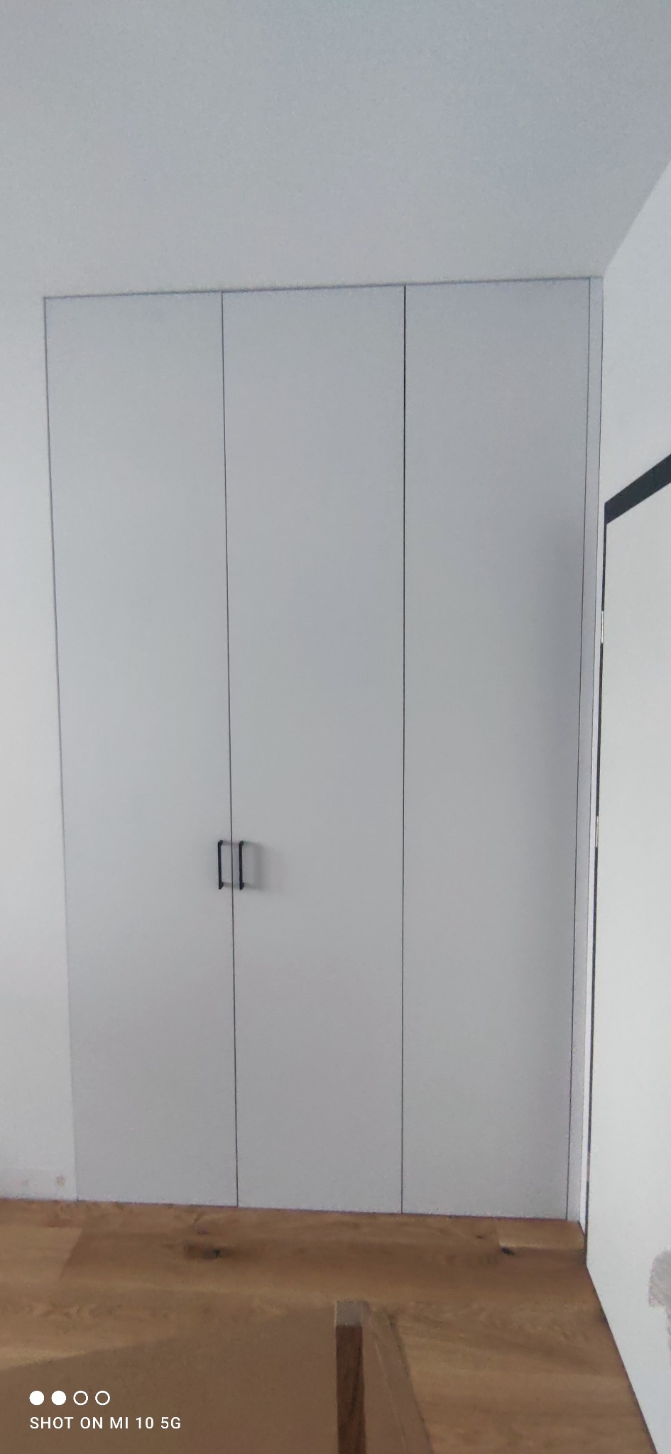 Trzydrzwiowa szafa wnękowa z szarymi frontami i minimalistycznymi czarnymi uchwytami, widoczna na tle drewnianej podłogi i białych ścian w trakcie wykańczania.