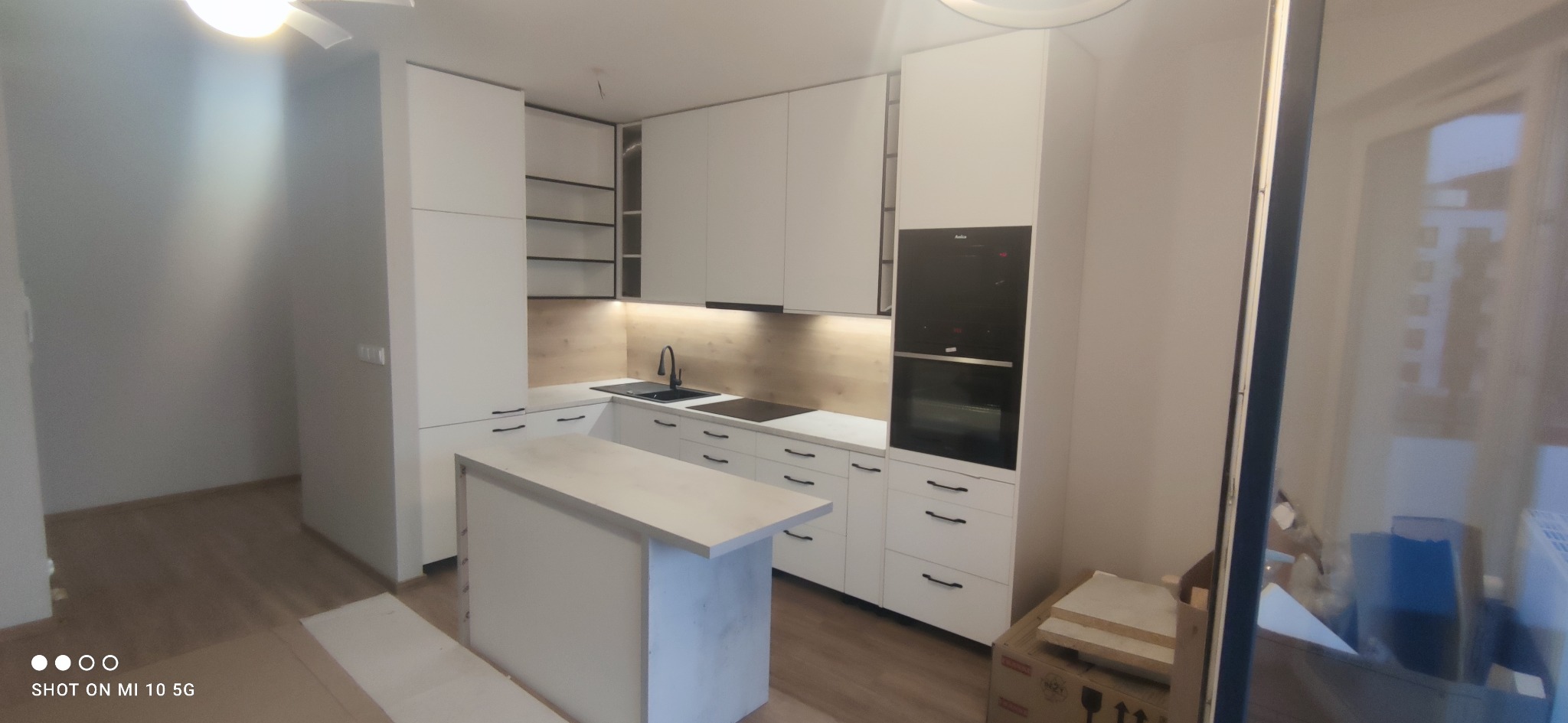 Nowoczesna biała kuchnia z wyspą, czarnymi akcentami i drewnianym panelem ściennym, widoczna część podłogi i kartony po transporcie mebli.