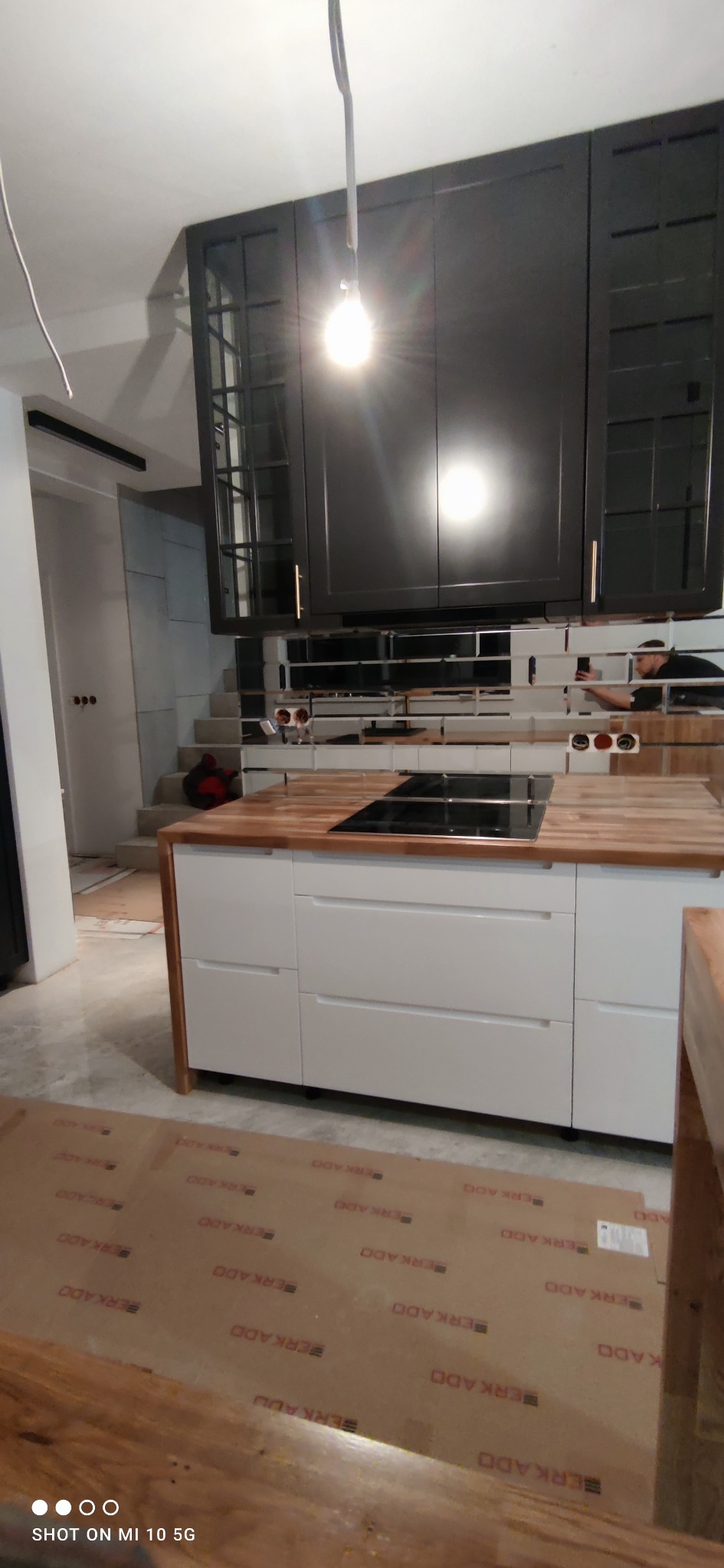 Montaż kuchni: czarne szafki wiszące z przeszklonymi frontami, drewniany blat z płytą indukcyjną, białe szafki dolne z szufladami, widoczny fragment schodów w tle, oświetlenie robocze włączone,...