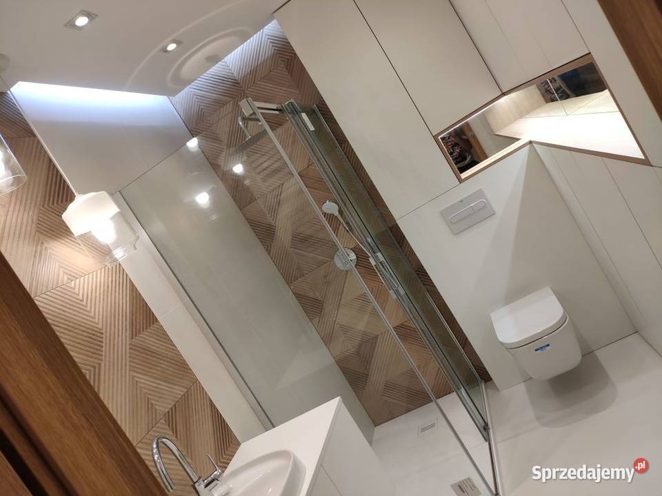 Nowoczesna łazienka z kabiną prysznicową, umywalką z baterią i podwieszaną toaletą, wykończona płytkami w geometryczny wzór drewna, oświetlona lampą wiszącą i wbudowanym oświetleniem sufitowym.