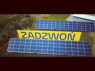 Dwa panele fotowoltaiczne ułożone obok żółtego billboardu z napisem 'ZADZWOŃ' w Ełku. Widok z góry na zielone otoczenie.