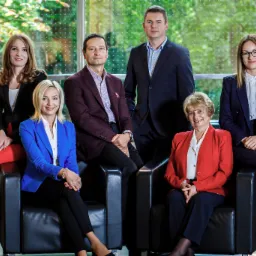 Zespołowe zdjęcie sześciu osób w eleganckich strojach siedzących w czarnych fotelach na tle okna z widokiem na zieleń.