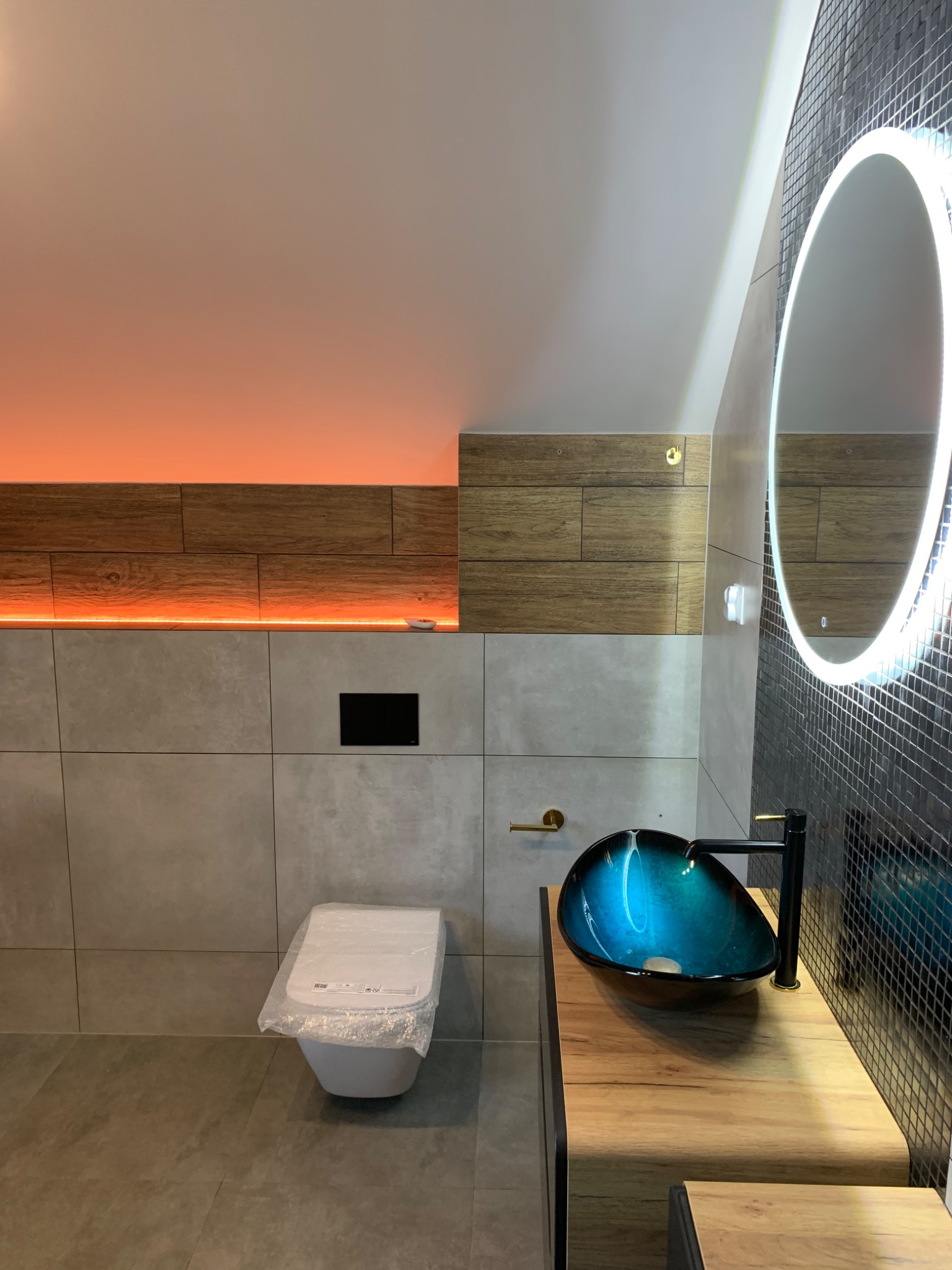 Nowoczesna łazienka z podświetlanym sufitem, umywalką w odcieniach niebieskiego i czarną armaturą, lustro z okrągłym podświetleniem LED, toaleta jeszcze w folii ochronnej.