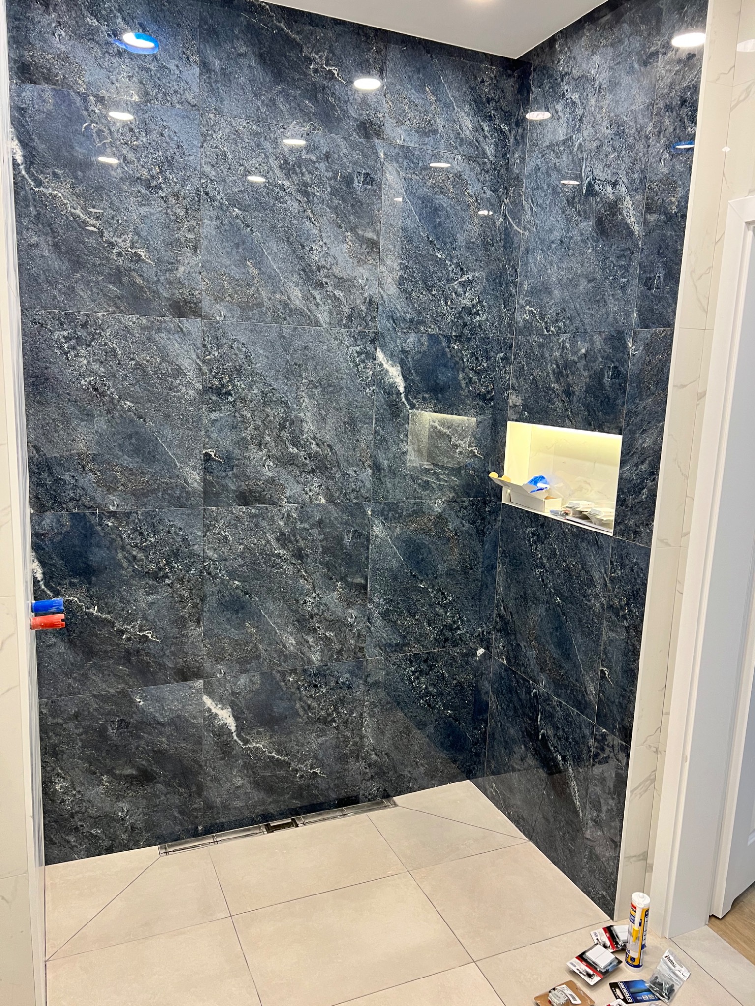 Wykończona kabina prysznicowa z ciemnymi, marmurkowymi płytkami na ścianach, jasnymi płytkami podłogowymi oraz wnęką z oświetleniem i akcesoriami. Na podłodze widoczne narzędzia i materiały montażowe.