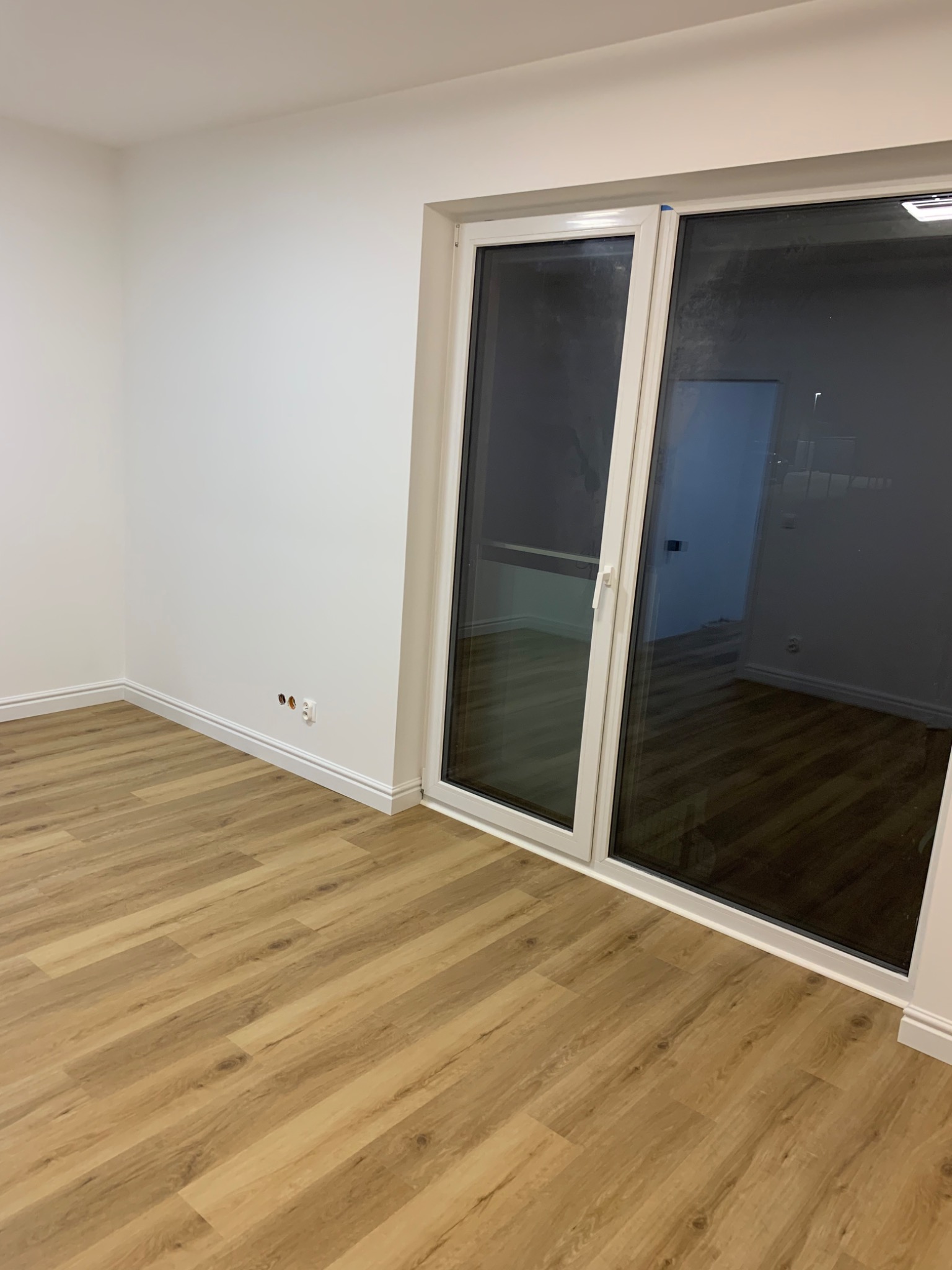 Pusty pokój z panelami podłogowymi w odcieniach drewna, białe ściany z listwami przypodłogowymi, gniazdka elektryczne i duże okno balkonowe z białymi ramami.