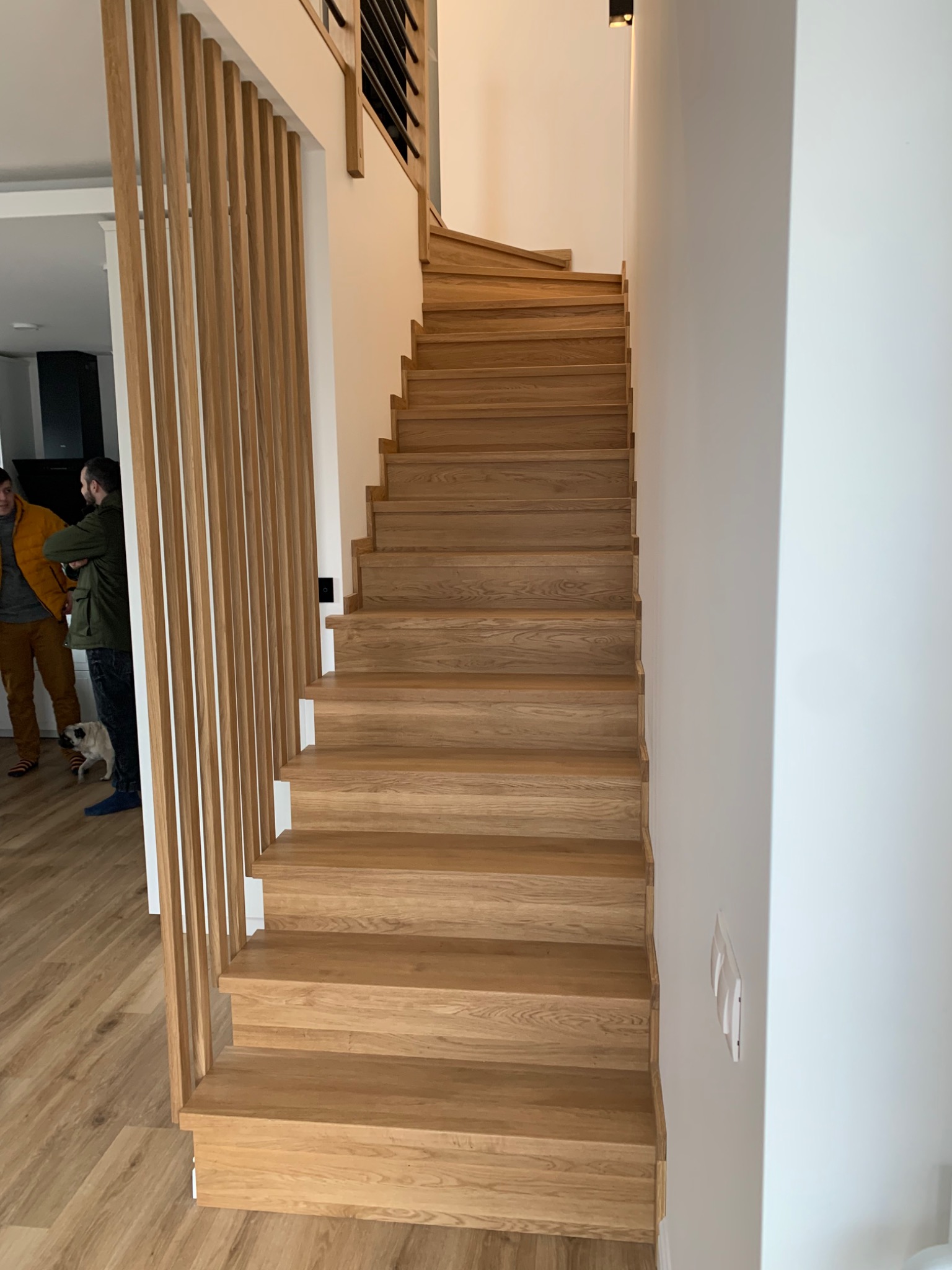 Nowoczesne schody drewniane z jasnego drewna, z pionowymi listwami oddzielającymi przestrzeń, widok z dołu, w tle dwie osoby i pies rasy pug.