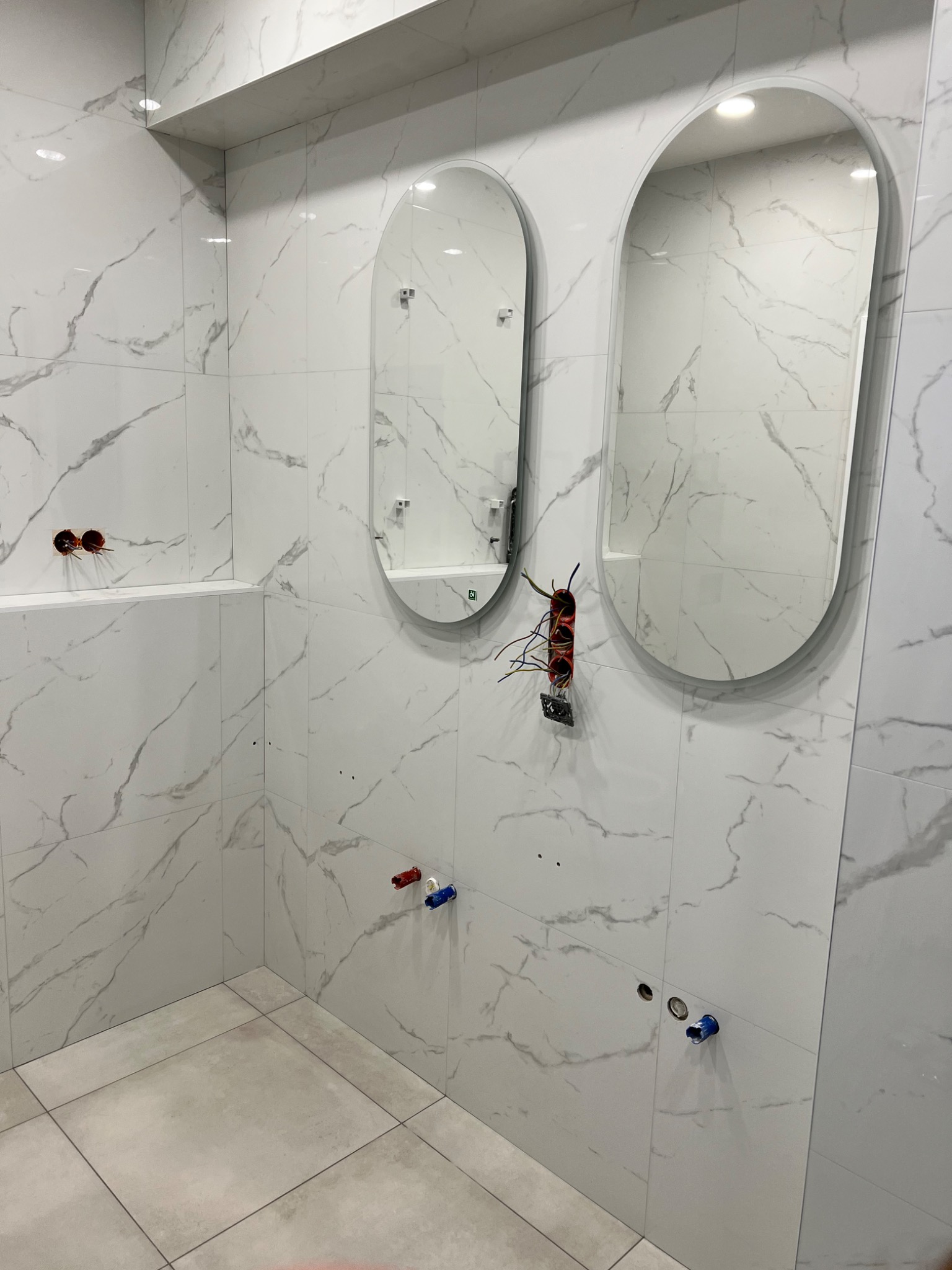 Łazienka w trakcie remontu: białe płytki imitujące marmur, dwa owalne lustra bez ram, wystające kable ze ściany, instalacja wodna w trakcie montażu.