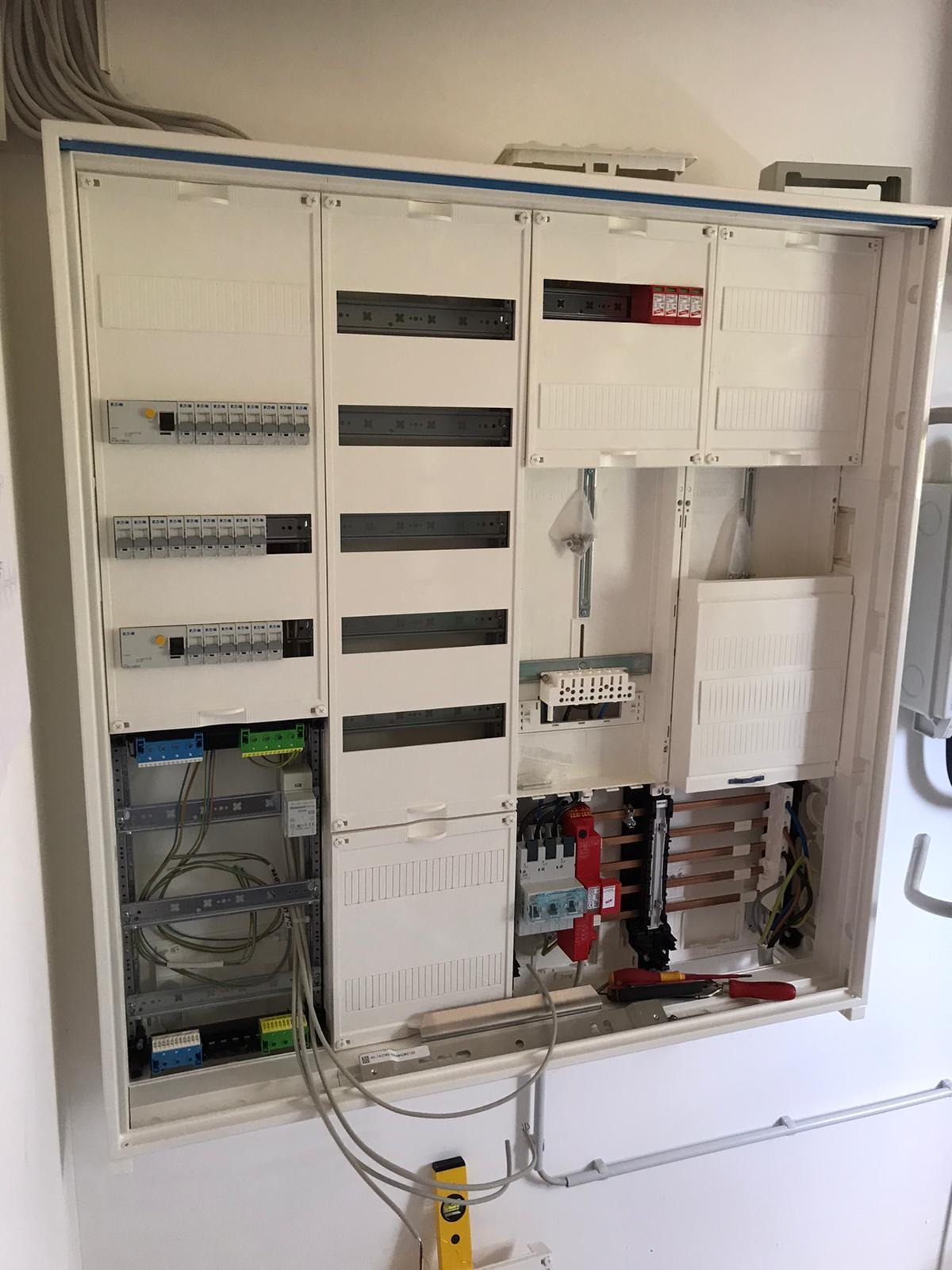 Otwarta, biała skrzynka rozdzielcza z widocznymi przewodami, bezpiecznikami i listwami zaciskowymi, poziomica na dole, przewody wychodzące ze skrzynki.