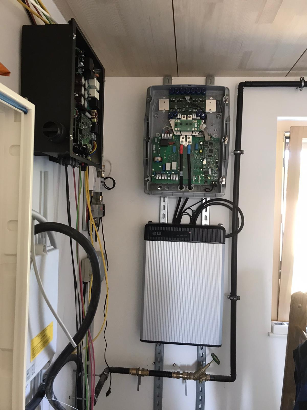 Komponenty instalacji fotowoltaicznej: otwarte skrzynki z elektroniką, akumulator LG na szynie montażowej, rury instalacyjne przy ścianie.