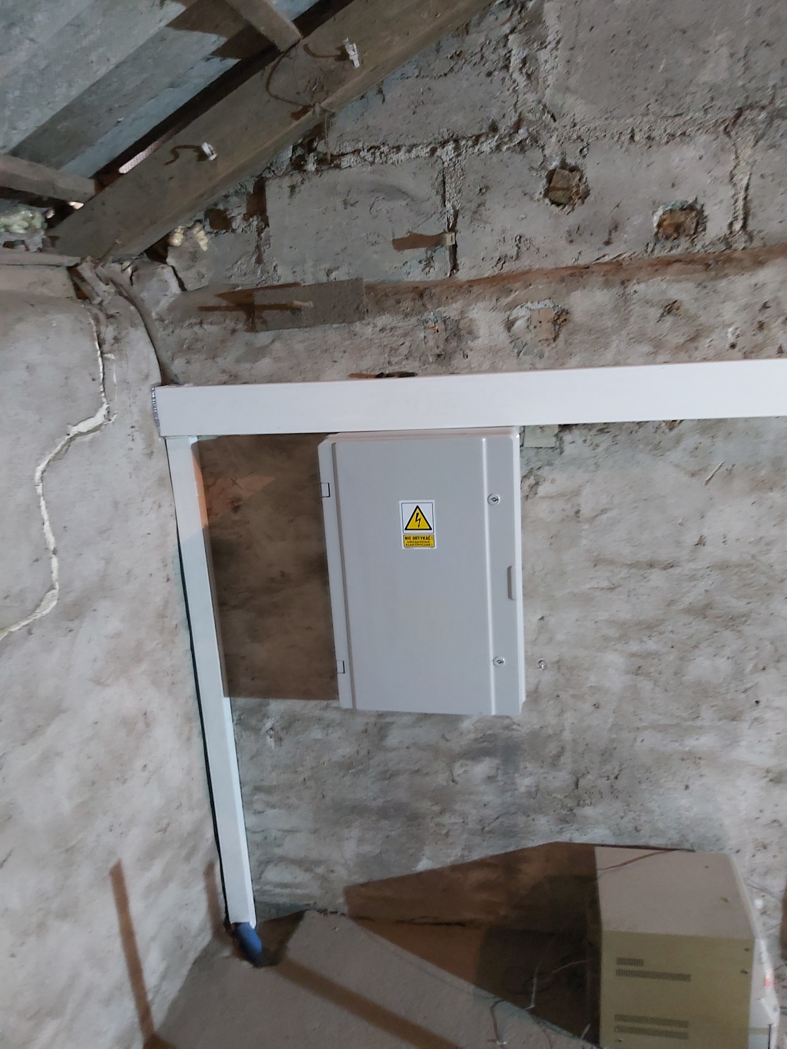 Szara skrzynka elektryczna z ostrzeżeniem o wysokim napięciu, zamontowana na surowej, nierównej ścianie w piwnicy, z widocznymi elementami instalacji i drewnianą konstrukcją stropu.