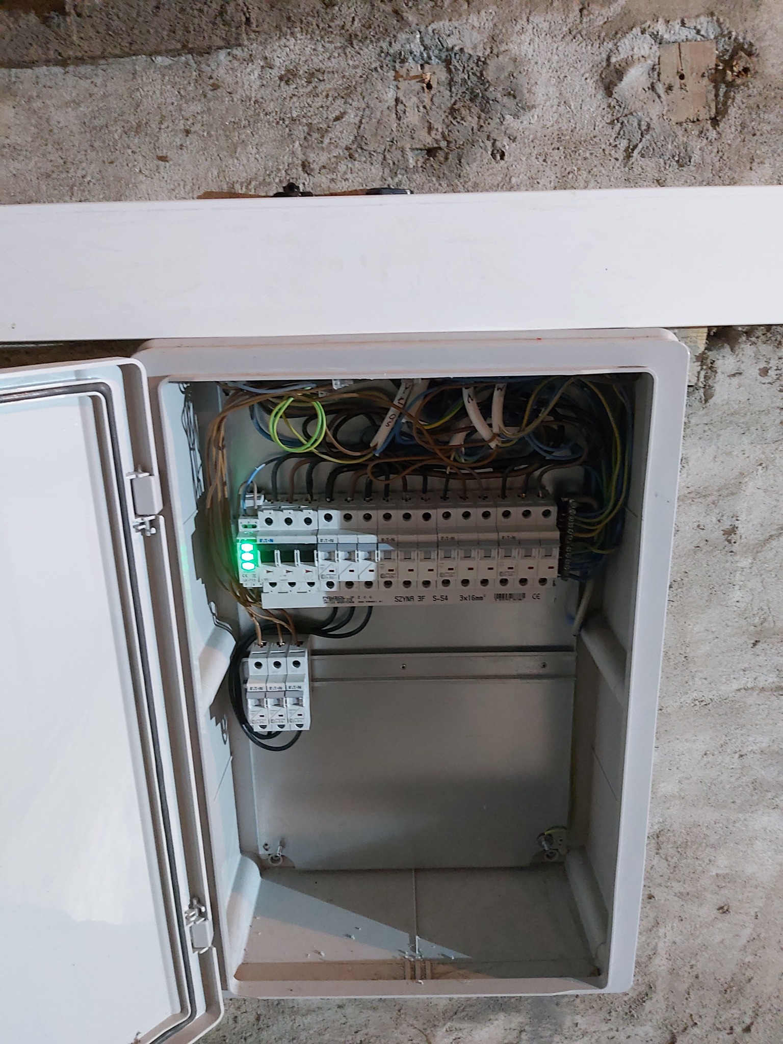 Otwarta skrzynka rozdzielcza z zainstalowanymi bezpiecznikami i przewodami, widoczne zielone diody sygnalizacyjne, tło stanowi surowa ściana.