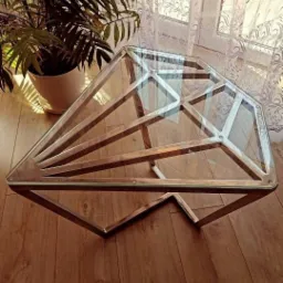 Szklany stolik kawowy o geometrycznym kształcie diamentu z metalową ramą, stojący na drewnianej podłodze w pobliżu okna z firanką i rośliną doniczkową.