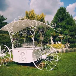 Ozdobny, biały wózek z metalowymi kołami i baldachimem, z dekoracyjnymi zawijasami, umieszczony na trawniku w ogrodzie z rzeźbą i starannie przyciętymi krzewami.