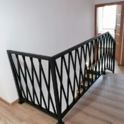 Czarna balustrada schodowa z geometrycznym wzorem krzyżujących się prętów, zamontowana w domu w Borzęcinie, widok z góry na drewnianą podłogę i białe ściany.