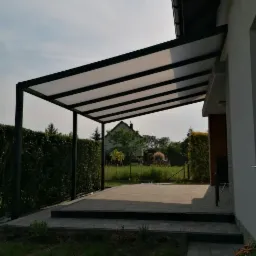 Zadaszenie tarasu z czarną metalową konstrukcją i mlecznym poliwęglanem, widok na ogród z trawnikiem i tujami w tle.