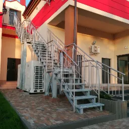 Zewnętrzne schody metalowe ze stalową balustradą prowadzące na piętro budynku z czerwonymi elementami architektonicznymi, obok klimatyzatory.