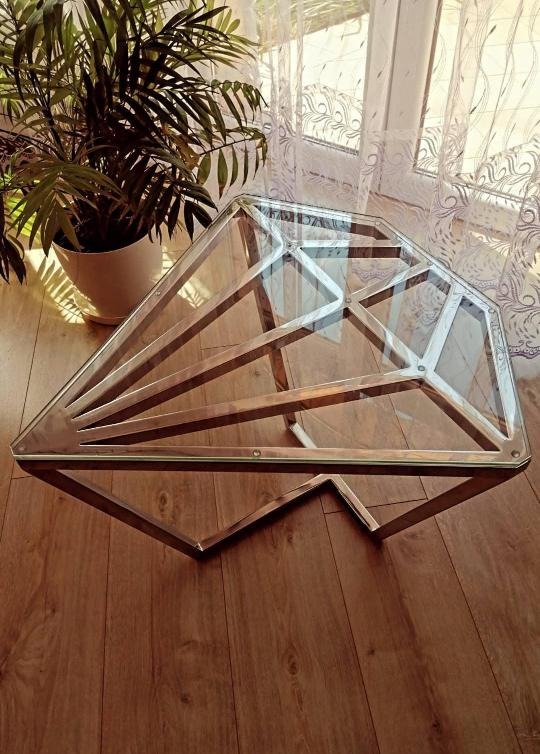 Szklany stolik kawowy o geometrycznym kształcie diamentu z metalową ramą, stojący na drewnianej podłodze w pobliżu okna z firanką i rośliną doniczkową.