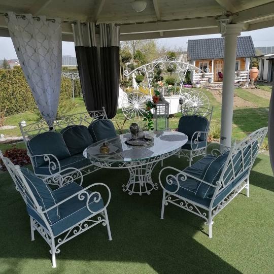 Komplet mebli ogrodowych z białego metalu z dekoracyjnymi elementami, składający się z sofy, dwóch foteli i krzeseł oraz owalnego stołu z szklanym blatem, ustawiony w altanie z zasłonami, w tle...