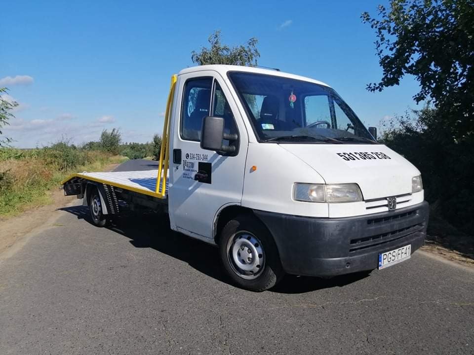 Biała laweta na drodze w Borku Wielkopolskim, widoczne numery kontaktowe na kabinie i żółte elementy platformy załadunkowej.