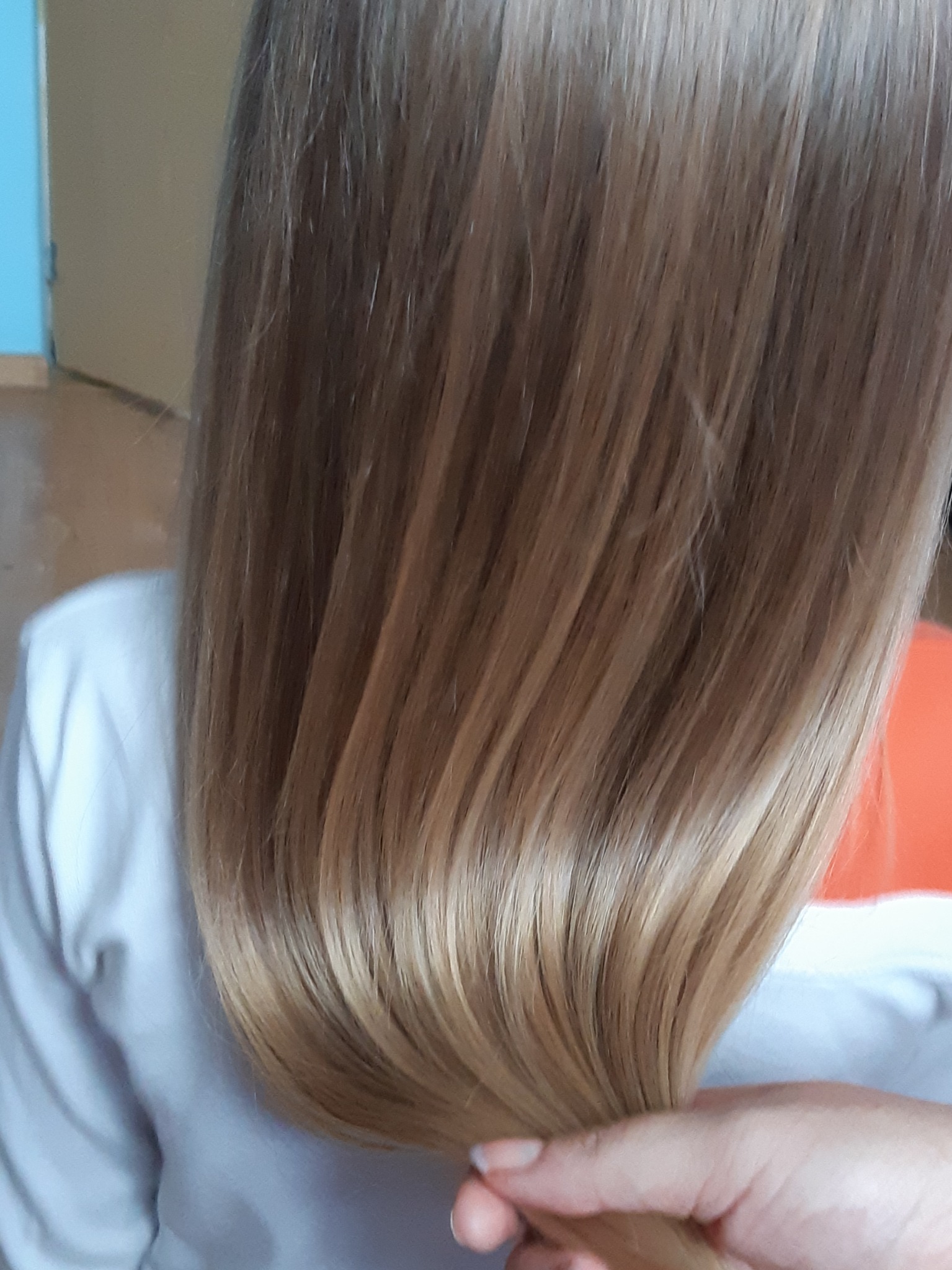 Proste, lśniące blond włosy z delikatnym ombre, trzymane w dłoni, ukazujące gładką strukturę po zabiegu pielęgnacyjnym.