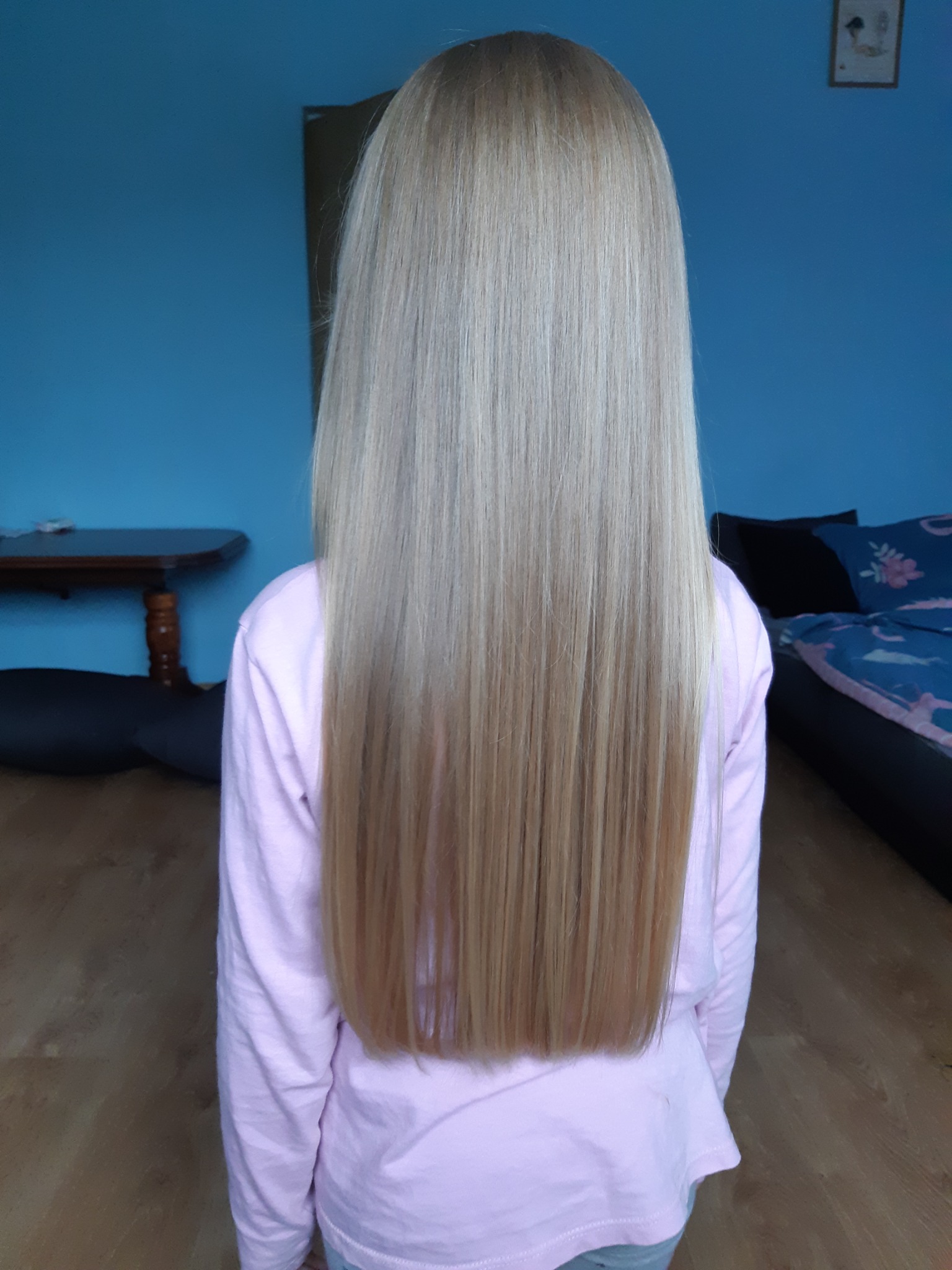 Długie, proste, blond włosy z delikatnym ombre, widok z tyłu na tle niebieskiej ściany i drewnianej podłogi.
