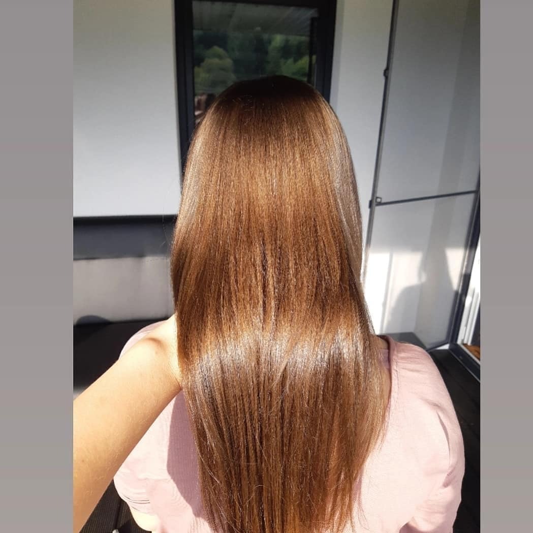 Długie, lśniące, kasztanowe włosy, widziane od tyłu, w naturalnym świetle, z widocznym cieniem na ścianie w tle.