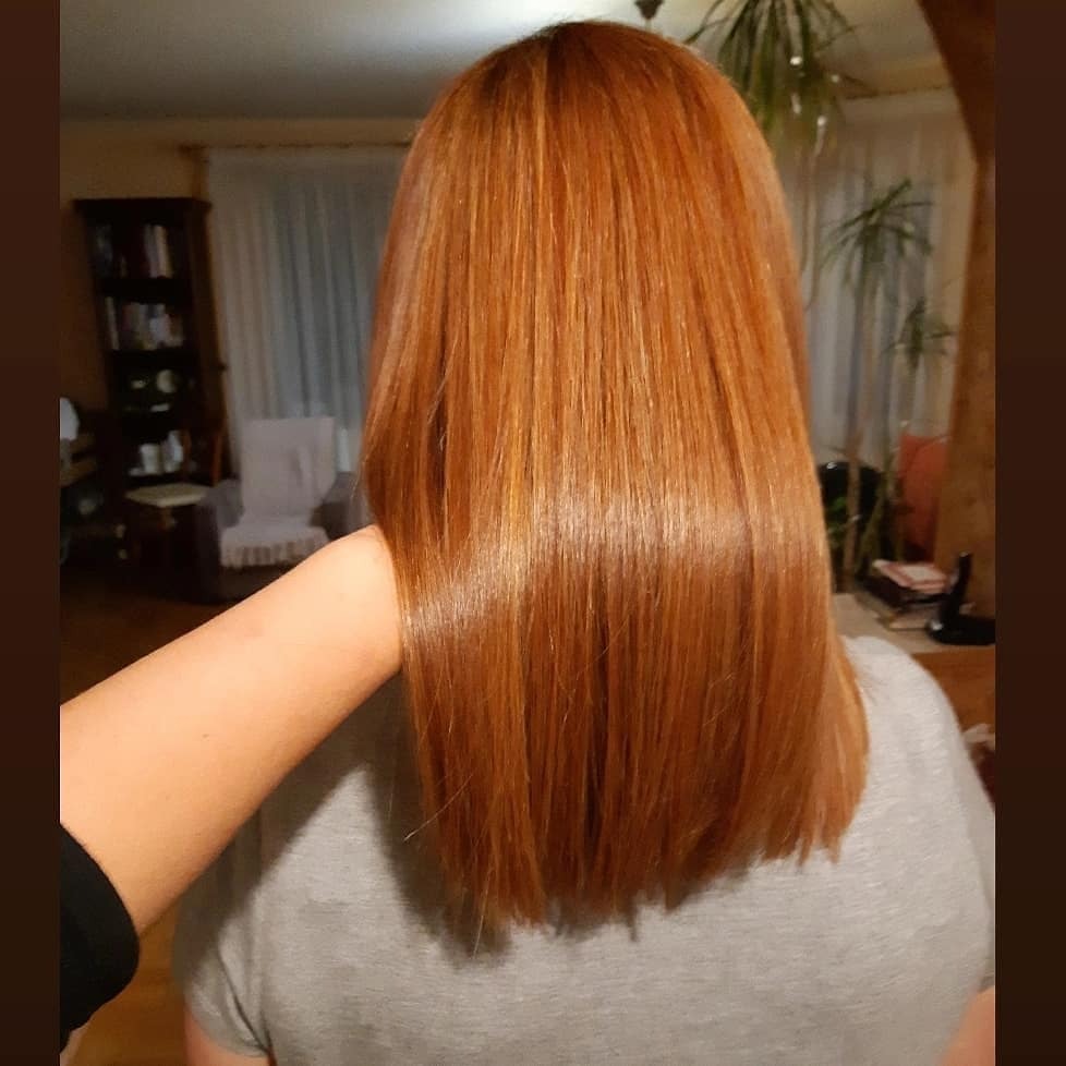 Gładkie, miedziane włosy po koloryzacji, widoczne z tyłu, lekko obcięte na prosto, z dłonią fryzjera delikatnie dotykającą pasma.