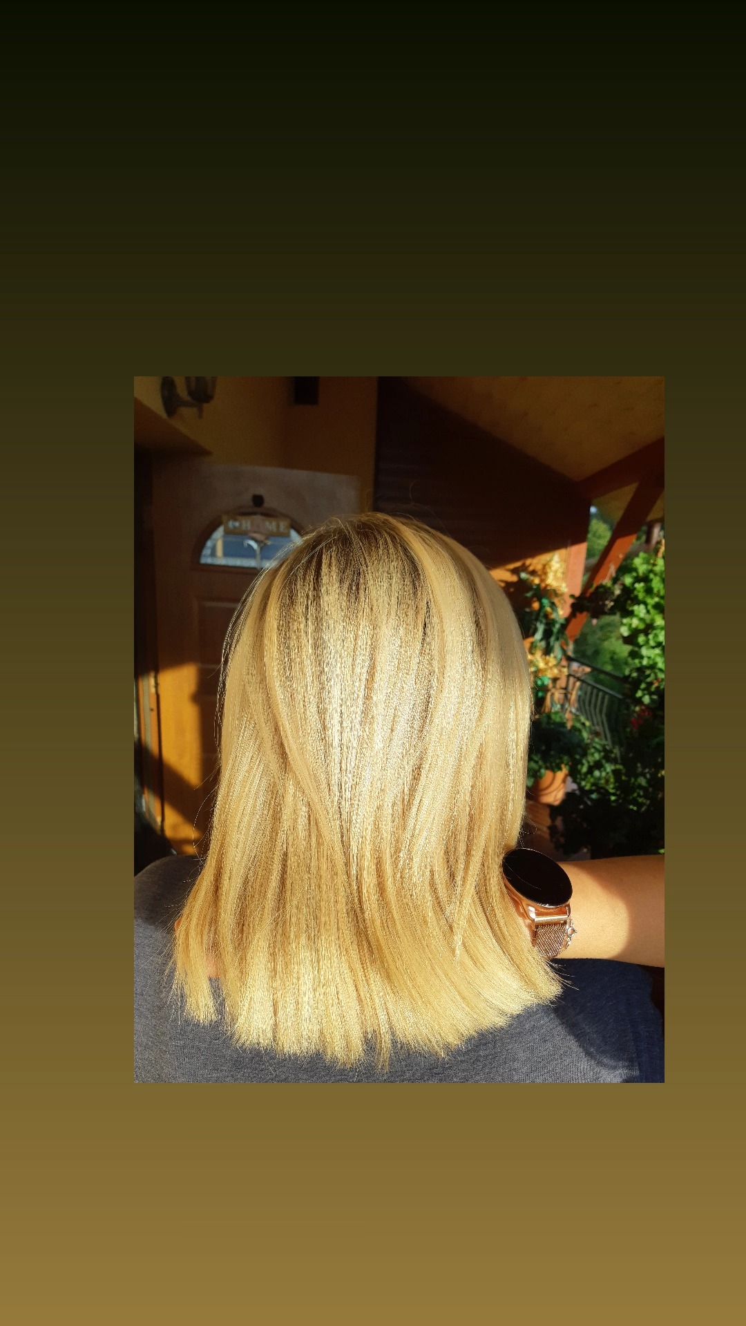 Tył głowy z włosami w odcieniach blondu, delikatnie cieniowane, z widocznym zegarkiem na ręce, w tle wejście do domu i zieleń.