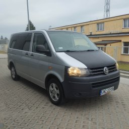 MATCARS POMOC DROGOWA Wynajem busów GIŻYCKO - Srebrny bus Volkswagen Transporter z czarną maską, zaparkowany na brukowanym placu, z budynkiem w tle. Widoczne tablice rejestracyjne i włączone światła.