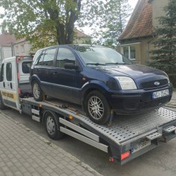 MATCARS POMOC DROGOWA Wynajem busów GIŻYCKO - Laweta z niebieskim Fordem Fusion na platformie, tablica rejestracyjna NGI 35425, w tle budynki mieszkalne i drzewa. Pomoc drogowa na osiedlowej ulicy.