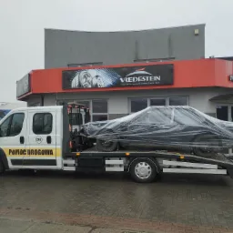 Laweta z uszkodzonym autem pod folią na tle budynku serwisu opon i samochodów. Pomoc drogowa 24h.