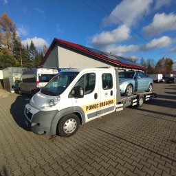 MATCARS POMOC DROGOWA Wynajem busów GIŻYCKO - Laweta z błękitnym autem osobowym na platformie, zaparkowana na brukowanym placu przed budynkiem z panelami słonecznymi na dachu, w słoneczny dzień.