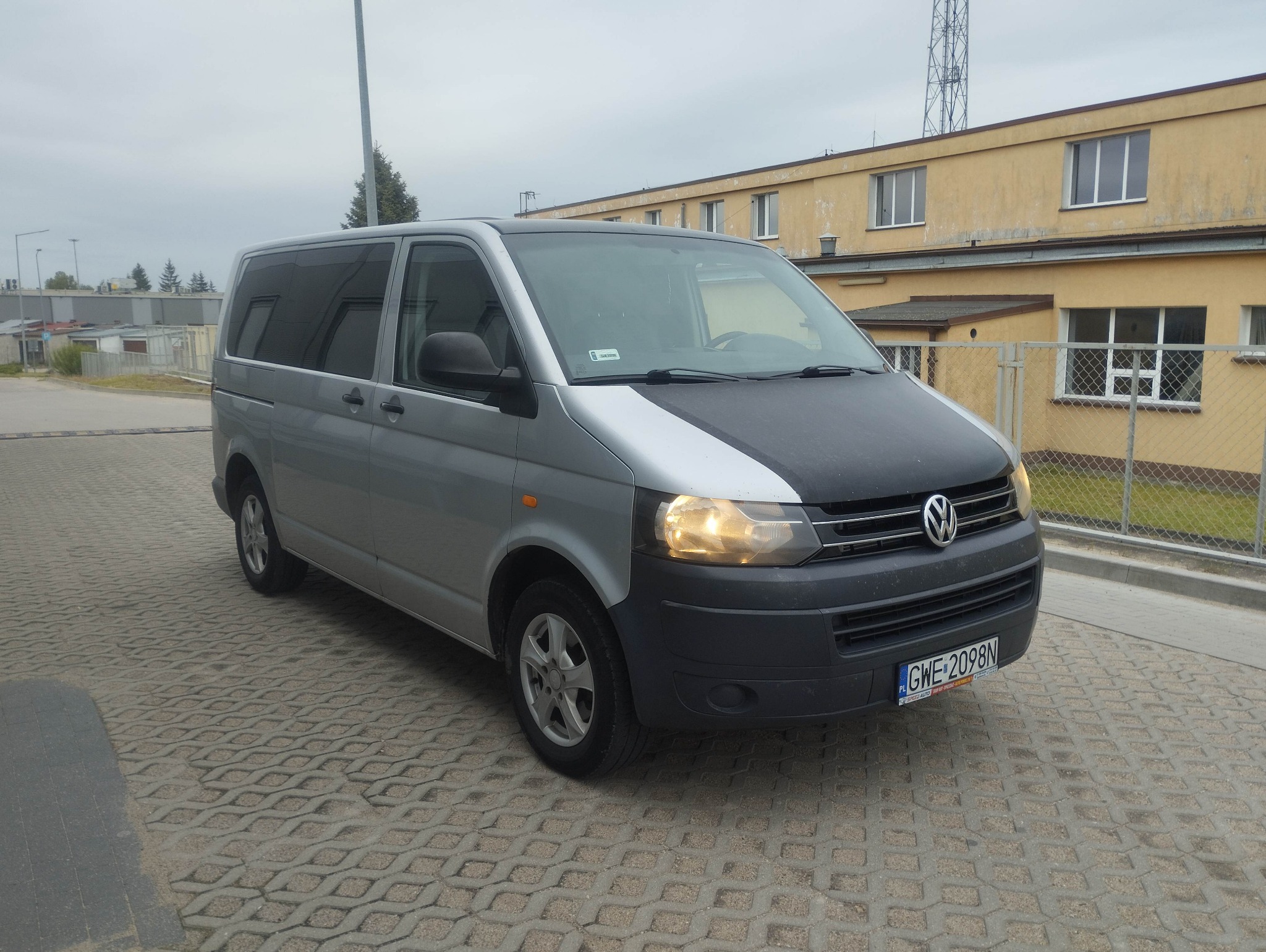 Srebrny bus Volkswagen Transporter z czarną maską, zaparkowany na brukowanym placu, z budynkiem w tle. Widoczne tablice rejestracyjne i włączone światła.